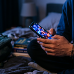 Teenager's hands holding a glowing smartphone in a dark bedroom, illustrating children's online safety debate