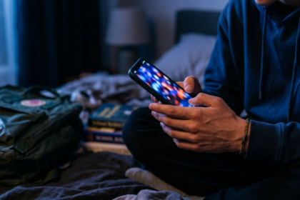 Teenager's hands holding a glowing smartphone in a dark bedroom, illustrating children's online safety debate